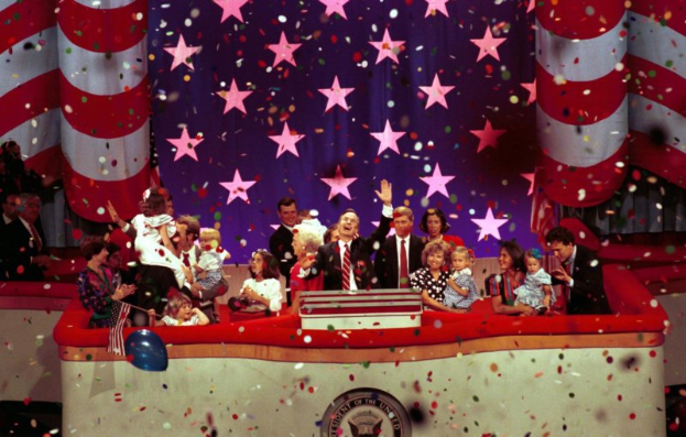 Eine Gruppe von Menschen steht auf einer Bühne mit einem Podium, das ein Logo trägt, umgeben von Luftballons und Konfetti, mit Sternen und einem Vorhang im Hintergrund.