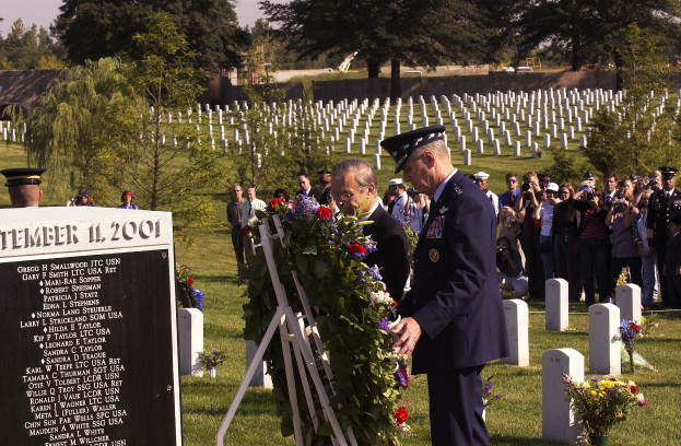 Eine Gruppe von Menschen steht um ein Grab auf einem Friedhof, während ein Mann in Uniform einen Kranz darauflegt, umgeben von Blumenbouquets, einer Tafel mit Text und Bäumen unter einem bewölkten Himmel.