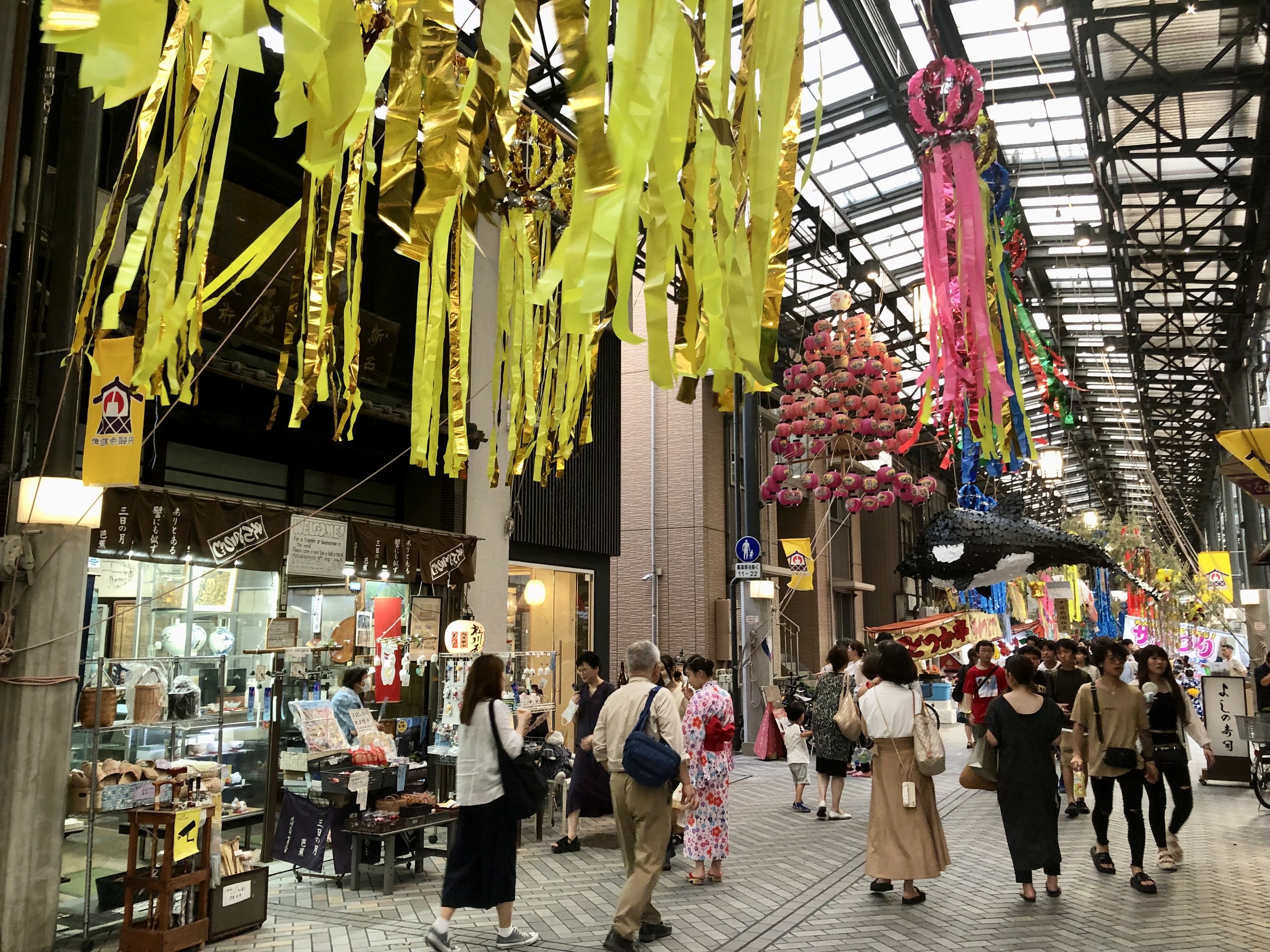 Menschen auf einer Straße mit Geschäften auf beiden Seiten, einige tragen Taschen, mit dekorativen Gegenständen an der Decke und einem Laden mit verschiedenen Artikeln links.