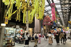 Menschen auf einer Straße mit Geschäften auf beiden Seiten, einige tragen Taschen, mit dekorativen Gegenständen an der Decke und einem Laden mit verschiedenen Artikeln links.