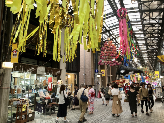 Menschen auf einer Straße mit Geschäften auf beiden Seiten, einige tragen Taschen, mit dekorativen Gegenständen an der Decke und einem Laden mit verschiedenen Artikeln links.