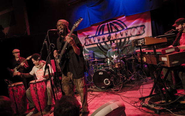 Eine Live-Band performt auf der Bühne mit Mikrofonen und Ständern, während das Publikum unten unter einer dunklen und mysteriösen Atmosphäre zusieht.