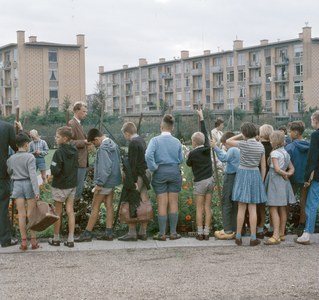 Kinder stehen vor einem Gebäude mit blühenden Pflanzen, Bäumen und einem klaren blauen Himmel, einige halten Schulranzen.
