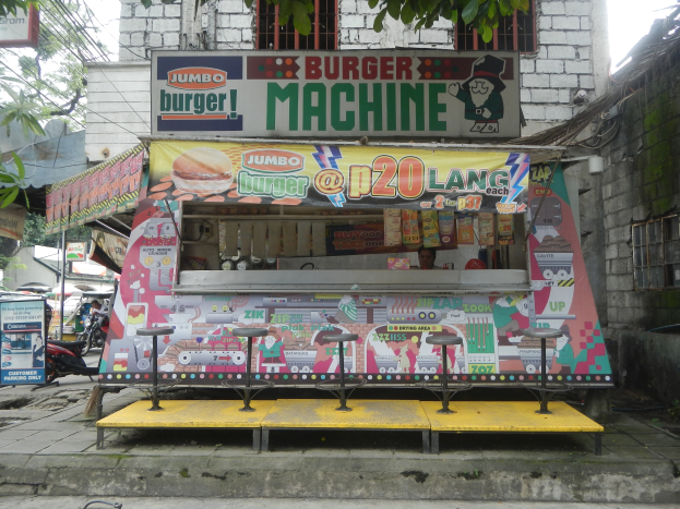 Ein bunt bemalter gelber und roter Foodtruck in Form eines Jumbo-Burgers, der auf einer Stadtstraße umgeben von Gebäuden, Bäumen und Menschen geparkt ist.