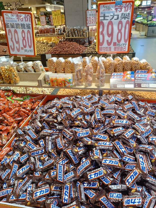 Vitrine mit verschiedenen Sorten von Süßigkeiten, einschließlich Snickers, in einem Geschäft mit Lebensmittelartikeln, nummerierten Schildern, Deckenleuchten und Hintergrundobjekten.
