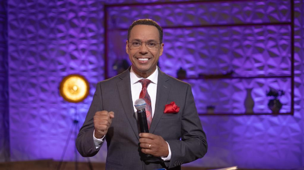 Stephen Colbert, Moderator von "The Daily Show with Trevor Noah", steht auf einer Bühne und hält ein Mikrofon, trägt einen Anzug, eine Krawatte und eine Brille, während hinter ihm ein Scheinwerfer und ein Ständer zu sehen sind.