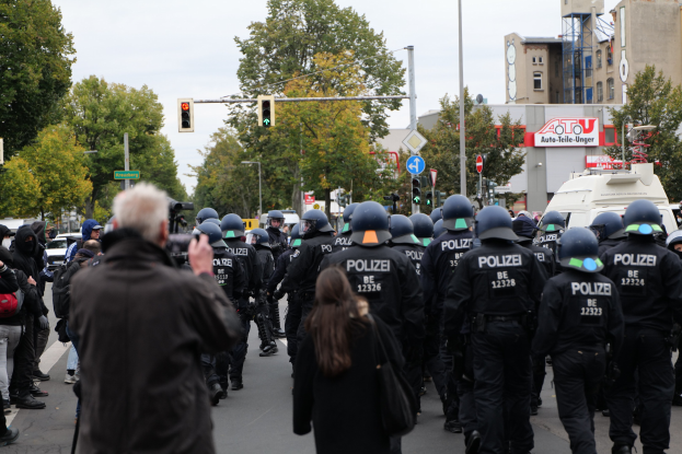 Eine Gruppe von Menschen geht eine Straße entlang mit Polizeibeamten, einige tragen Helme und tragen Taschen oder Kameras, mit Fahrzeugen, Verkehrszeichen, Schildern, Bäumen, Gebäuden und einem klaren Himmel im Hintergrund.