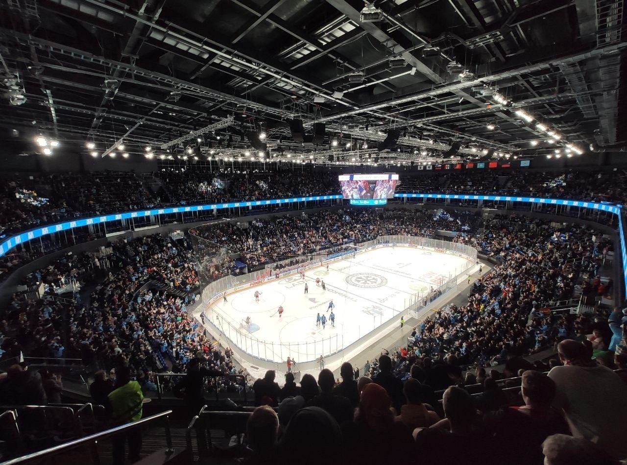 Eishockeyspiel in einer großen Arena mit Zuschauern auf den Tribünen, Geländern, Überbildschirm und heller Beleuchtung; als O2 Arena in London identifiziert.