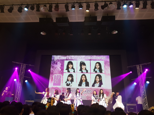 Eine Gruppe von Menschen performs auf einer hell erleuchteten Bühne mit Trägern, während ein Sänger ein Mikrofon hält und ein Musiker ein Instrument spielt, während eine Menge darunter die Veranstaltung aufzeichnet und ein großer Bildschirm Bilder der K-Pop-Gruppe BK48 anzeigt.