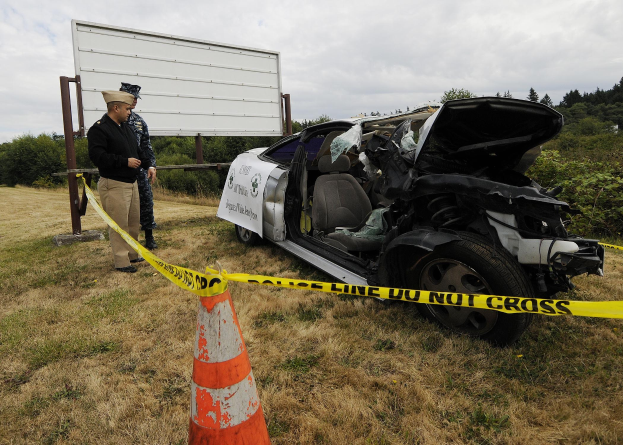 Ein Polizeibeamter in Uniform steht neben einem schwer beschädigten Auto am Straßenrand, mit eingedellter Motorhaube und zersplitterten Fenstern, neben einem Verkehrskegel und einer Schnur an einem Pfahl, mit Bäumen und einem bewölkten Himmel im Hintergrund.