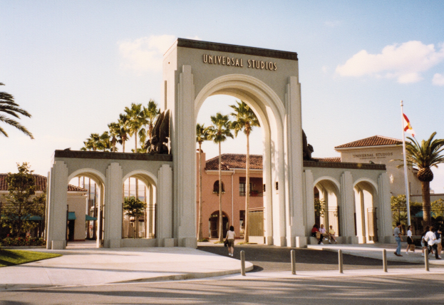 Eingang zu Universal Studios Hollywood mit einem Bogen, einer Statue, Menschen, Bäumen, Gebäuden, einem Fahnenmast mit einer Flagge und einem bewölkten Himmel.