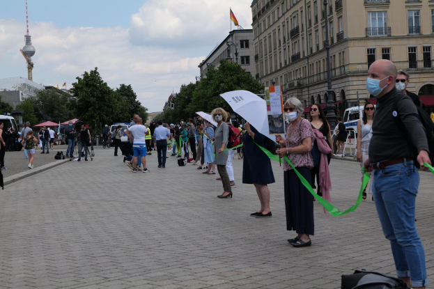 Eine Gruppe von Menschen, die eine Straße in Berlin, Deutschland, entlanggehen und Schilder und Banner halten, einige tragen Masken und tragen Taschen oder Schirme, mit Bäumen, Gebäuden, geparkten Fahrzeugen und einem Turm mit Flaggen im Hintergrund unter einem bewölkten Himmel.