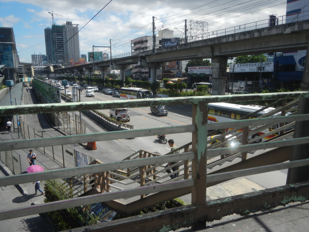 Eine belebte Stra├če in der Stadt von einer ├Überquerung aus betrachtet, mit Fahrzeugen, Passanten, Gel├ädern, Vegetation, Geb├Ąuden, Strommasten mit Kabeln.