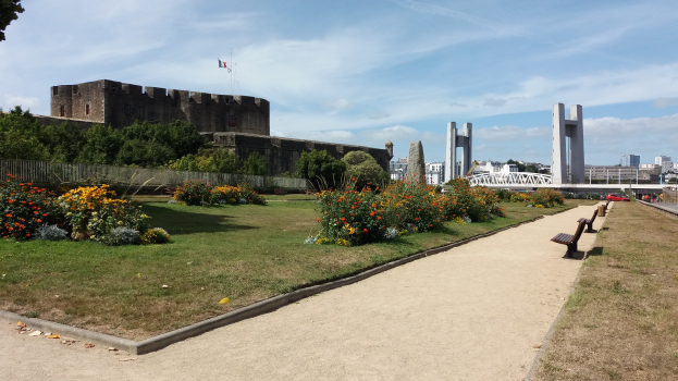Ein Park mit Bänken und Blumen vor einem Schloss, umgeben von Gras, blühenden Pflanzen, einem Zaun, Bäumen, Fahrzeugen, einer Brücke, Säulen, einem Fahnenmast mit einer Fahne, Gebäuden und einem bewölkten Himmel.