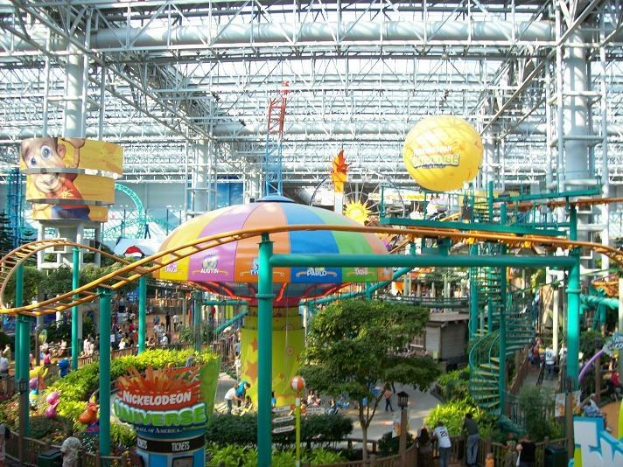 Indoor-Wasserpark mit einer Achterbahn umgeben von Menschen, Bäumen, Pflanzen und Deckenbeleuchtung mit Stangen.