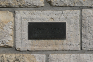 Steinwand mit einem rechteckigen schwarzen Schild, auf dem "National Youth Administration" in weißer, fetter, zentrierter Schrift steht.
