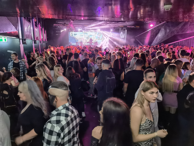 Eine große Gruppe von Menschen in einer Diskothek mit Deckenbeleuchtung und Säulen, mit einem Bildschirm im Hintergrund bei einem Absolut Nightlife-Event.
