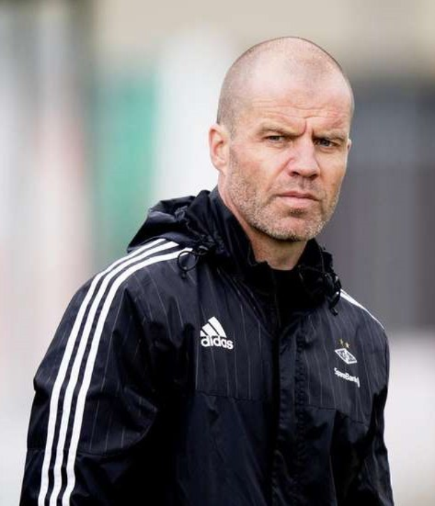 Ein kahlkopfeter Mann in einer schwarzen Jacke steht auf einem Fussballfeld, trägt eine marineblaue Jacke mit weißen Streifen und hat die Hände verschränkt, mit einem ernsten Gesichtsausdruck und einem unscharfen Hintergrund.