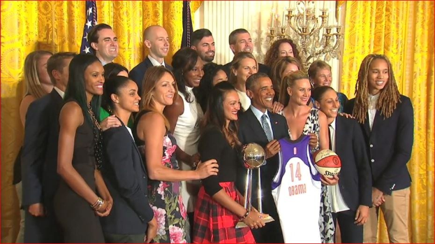 Präsident Obama und First Lady Michelle Obama posieren mit dem Frauen-Basketball-Team im Oval Office des Weißen Hauses, halten einen Basketball, eine Trophäe und lächeln neben einer Flagge, Vorhängen und einem Kerzenleuchter.