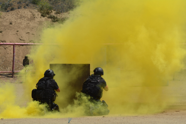 Zwei Polizisten in schwarzen Uniformen und Helmen lassen gelben Rauch in der Nähe eines Metallzauns mit Sand, Pflanzen und Bäumen im Hintergrund frei.