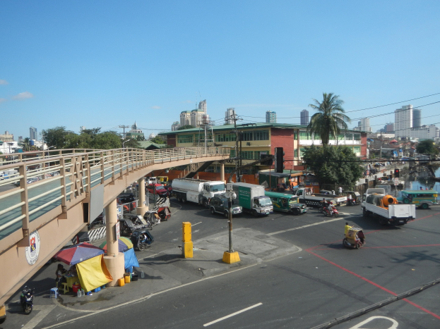 Vielbefahrene Straße mit Schwerverkehr, Föülern, Zelten und urbanen Elementen neben einer überquerung über Wasser unter einem bewölkten Himmel.