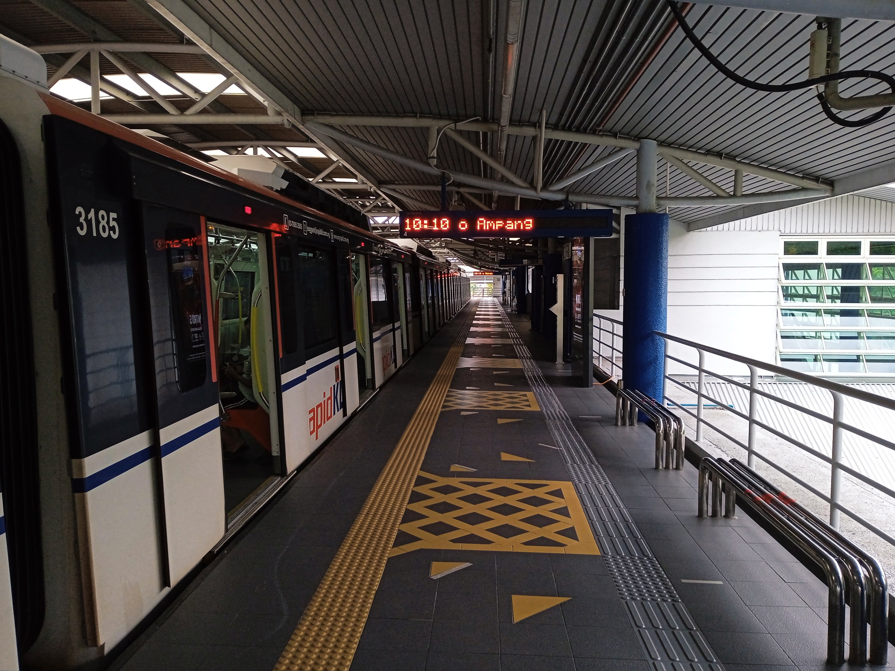 Ein Zug fährt in einen Bahnhof ein, neben einem Bahnsteig mit Geländern, Säulen und einer Tafel mit Text, mit Glasfenstern und einer Stadtlandschaft im Hintergrund.