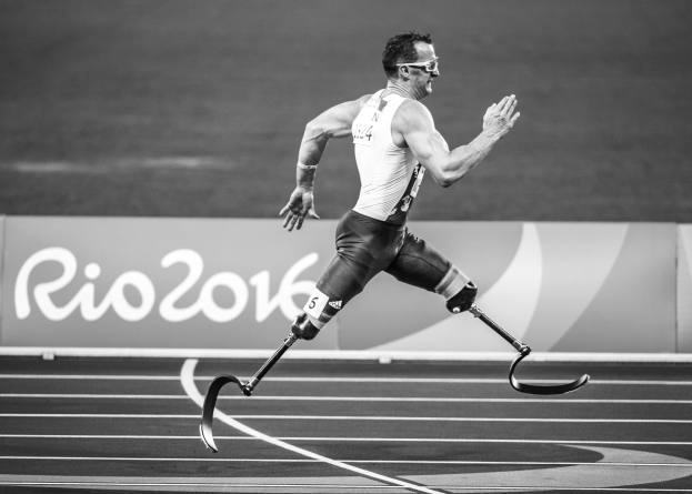 Ein Mann mit einer Prothese rennt auf einer Laufbahn mit einer Tafel "Rio 2016 Paralympische Spiele" im Hintergrund, in Schwarz-Weiß.