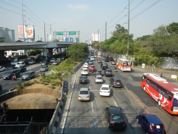 Eine volle städtische Straße mit Fahrzeugen, Oberleitungen, Bäumen, Gebäuden, Werbetafeln und einer fernen Brücke unter einem bewölkten Himmel.