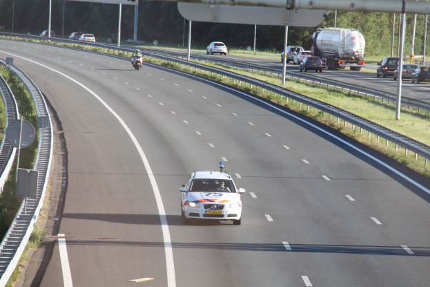 Ein Auto auf einer Autobahn mit einem stationären Polizeiwagen am Straßenrand, umgeben von anderen Fahrzeugen, Leitplanken, Strommasten, Schildern und Bäumen.