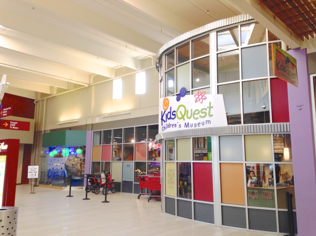 Innenansicht eines Einkaufszentrums mit einem "Kids Quest Kindermuseum"-Schild, Einkaufswagen, ribbonierten Pfählen, Texttafeln, Deckenleuchten und verstreuten Gegenständen.
