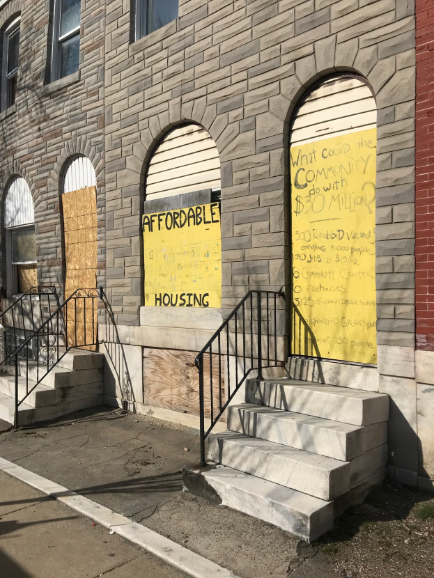 Ein zweistöckiges Gebäude mit vernagelten Fenstern und einem Schild mit der Aufschrift "Günstiger Wohnraum", das über eine Treppe mit Geländern erreichbar ist und neben einer Straße steht.