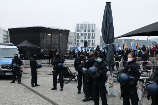 Polizeibeamte in Uniform hinter einer Absperrung vor einer Menge von Demonstranten, einige mit Helmen und Masken, mit Zelten, Fahnen und Gebäuden im Hintergrund bei einer Demonstration in Berlin gegen Regierungssparmaßnahmen.
