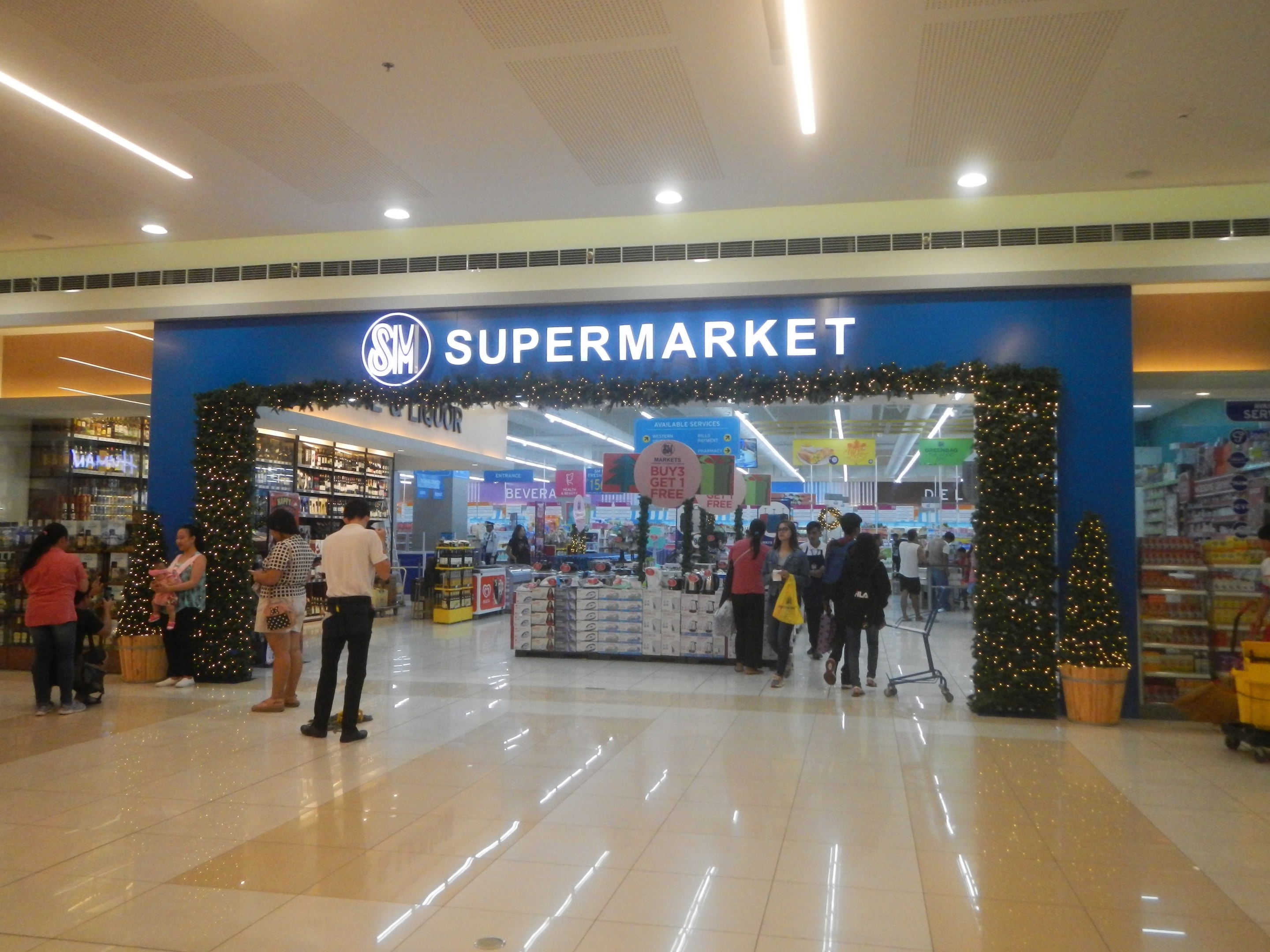 Eine Gruppe von Menschen vor einem Supermarkt, einige halten Taschen, mit einem Einkaufswagen, einem Topf mit Pflanze, Regalen, Schildern und einer Decke mit Deckenbeleuchtung.