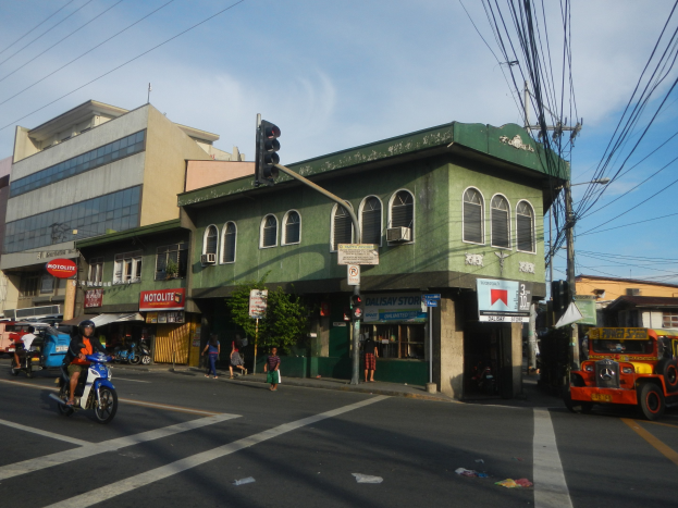 Eine belebte Stadtstraße mit einem grünen Gebäude im Hintergrund, Fahrzeugen auf der Straße, Fußgängern, einer Ampel an einem Mast, bewölktem Himmel und Strommasten mit Überlandleitungen.