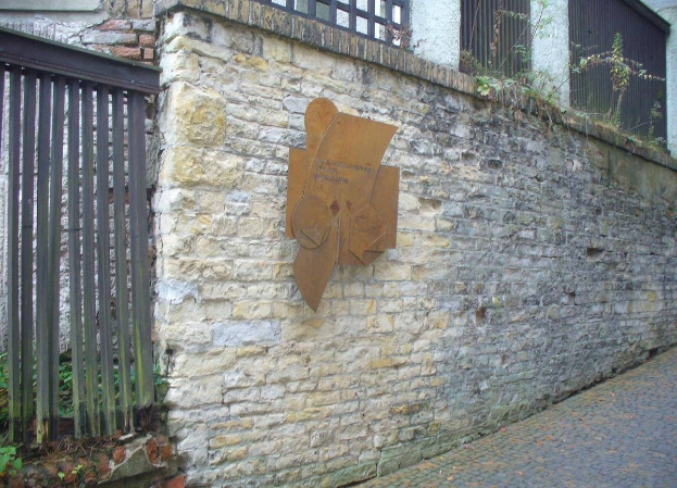 Steinmauer mit Metalltor auf der linken Seite, eine Tafel zur Einweihung des alten Rathauses und ein Gebäude mit Fenstern und Pflanzen im Hintergrund.