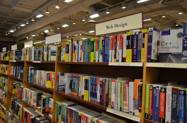 Eine Bibliothek mit Bücherregalen voller Bücher, Informationsplakate an den Wänden, Deckenbeleuchtung und sichtbare Rohre an der Decke.