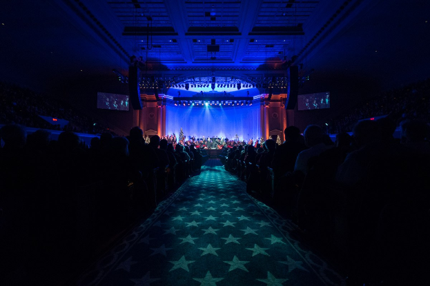 Ein großer Saal mit sitzenden Zuschauern, eine Bühne mit Musikern und eine Decke mit Lichtern und Fernsehgeräten, was auf ein Konzerthaus während der Nationalkonvention 2019 hindeutet.