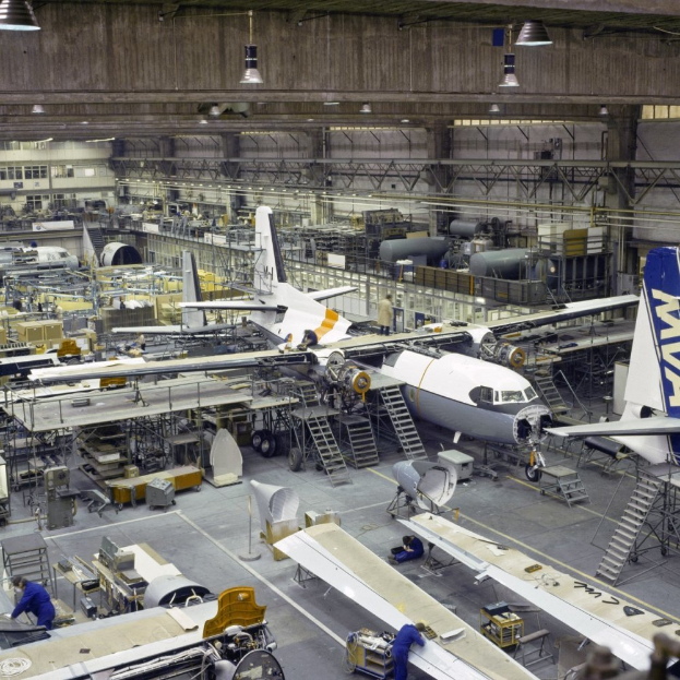 Ein großer Hangar voller Flugzeuge auf der Montagelinie, Arbeiter, Leitern und verschiedene Gegenstände auf dem Boden, mit Lampen an der Decke und einer Wand im Hintergrund.