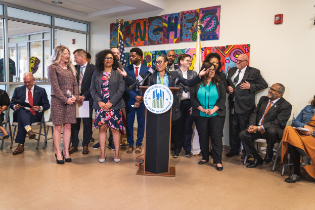 Eine Gruppe von Menschen steht vor einem Podium mit einem Mikrofon und einer Tafel mit der Aufschrift "Sancho-Stadtrat tritt dem Stadtrat bei", während andere sitzen oder in der Nähe stehen in einem Raum mit Flaggen, einem Feuerlöscher und Deckenleuchten.