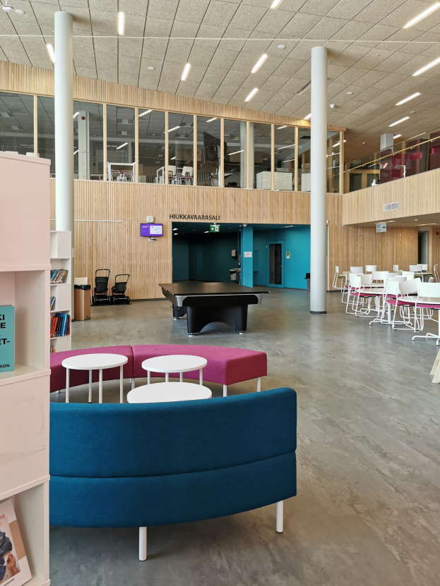 Großer Raum mit mehreren Tischen, Stühlen, Sofas, Hockern, Regalen, Säulen, Deckenleuchten und einem wandmontierten Fernseher, der an ein Schulbibliothek-Interieur erinnert.