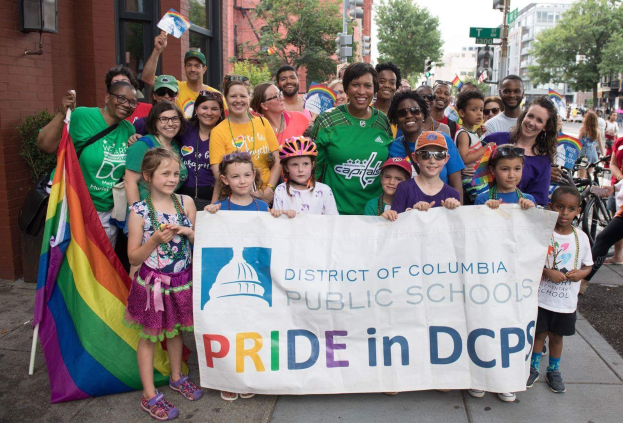 Eine Gruppe von Menschen, die draußen stehen, einige halten ein Banner mit der Aufschrift "District of Columbia Public Schools Pride in DCPS", mit Kindern, die eine Fahne halten, einem Radfahrer, Bäumen, Gebäuden, Schildern, Straßenlaternen und einem bewölkten Himmel.