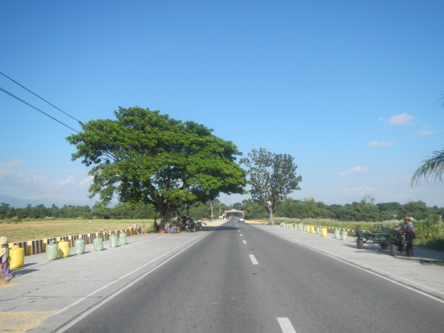 Weiße Straßenmarkierungen mit Motorradfahrern, Fußgängern am Straßenrand, Containern, einem Zaun, Strommasten, einer Oberleitung, Bäumen, Gras, fernen Hügeln und einem klaren blauen Himmel.