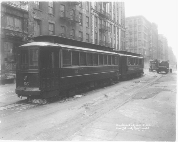 Ein Schwarz-Weiß-Foto eines Straßenbahnwagens auf einer Stadtstraße, umgeben von Gebäuden mit Fenstern, Treppen mit Geländern und Fahrzeugen auf der Straße, mit Text unten.