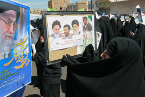 Eine Gruppe von Frauen in schwarzen Abayas hält Plakate mit Fotos von Ayatollah Ali Khamenei hoch, mit Gebäuden, Pfählen, Fahrzeugen und einem klaren blauen Himmel im Hintergrund.