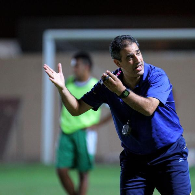 Ein Mann in einem blauen T-Shirt und Hose steht auf einem Fußballfeld und hält ein Mikrofon in der Hand, während er auf Spieler zeigt. Im Hintergrund sind eine Person in einem grünen Kleid, ein Torpfosten und eine Wand zu sehen.