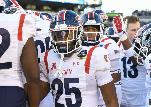 Gruppe von Männern in Fußballtrikots auf einem Feld mit Helmen und Handschuhen, Hintergrund zeigt unscharfe Zuschauer, ein Scoreboard mit "Auburn vs Virginia Tech - 11/28/15 College Football Pick, Odds, and Prediction" und Himmel.