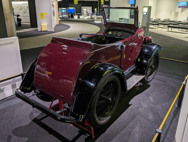 Ein rotes Oldtimer-Auto in einem Museum ausgestellt mit Informationsschildern, Barrieren und Sitzgelegenheiten.