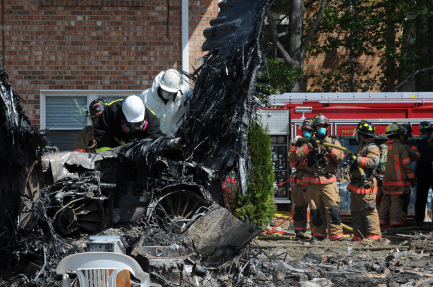 Feuerwehrleute in Schutzausrüstung löschen ein Gebäude Feuer, mit verkohlten Überresten einschließlich eines Fahrzeugs, eines Stuhls und Pflanzen auf dem Boden.