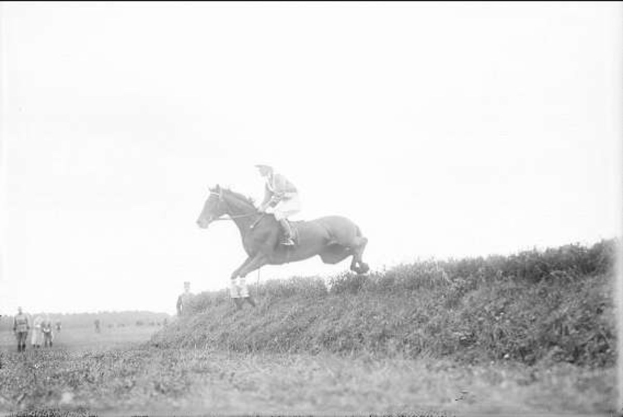 Schwarzes und weißes Foto eines Pferdes und Reiters, die über einen grünen Hügel springen, mit dem Pferd im Galopp und dem Reiter mit Helm und Jacke, während Zuschauer im Hintergrund beobachten.
