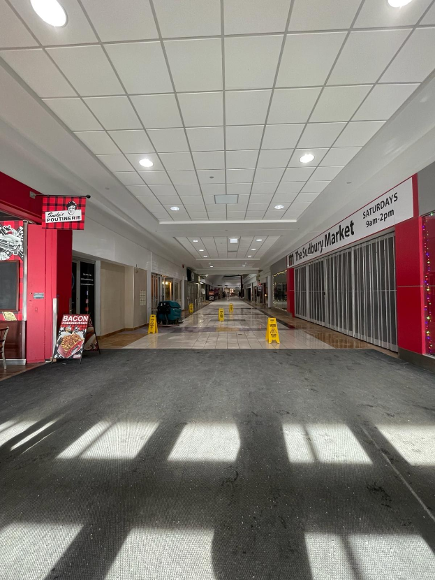 Innenansicht eines Einkaufszentrums mit Deckenleuchten, einem Stuhl und einer Tafel, auf der links "Sydbury Market - Vorher und Nachher" steht, sowie verstreuten Gegenständen rechts und auf dem Boden.